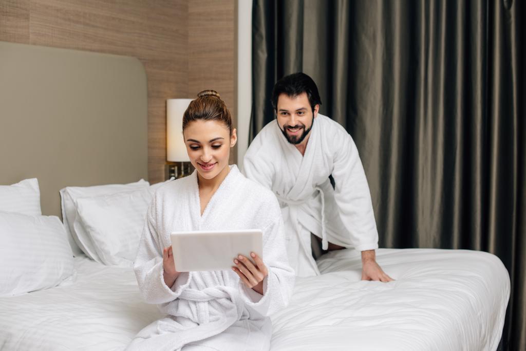 バスローブのタブレットを使用して遊び心のある彼氏が彼女に後ろからこっそりながらホテルのスイート ルームのベッドの上で美しい女性 ロイヤリティフリー 写真 画像素材