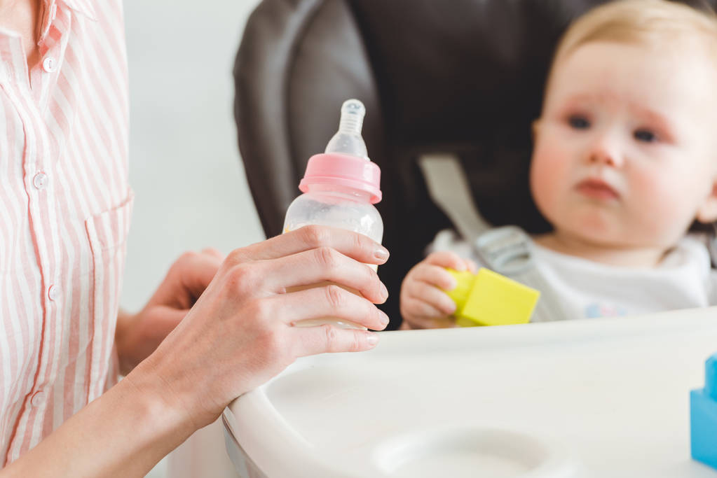 哺乳瓶とプラスチック製のブロックで赤ちゃん用の椅子に座っている幼い娘を持つお母さんのビューをトリミング ロイヤリティフリー写真 画像素材