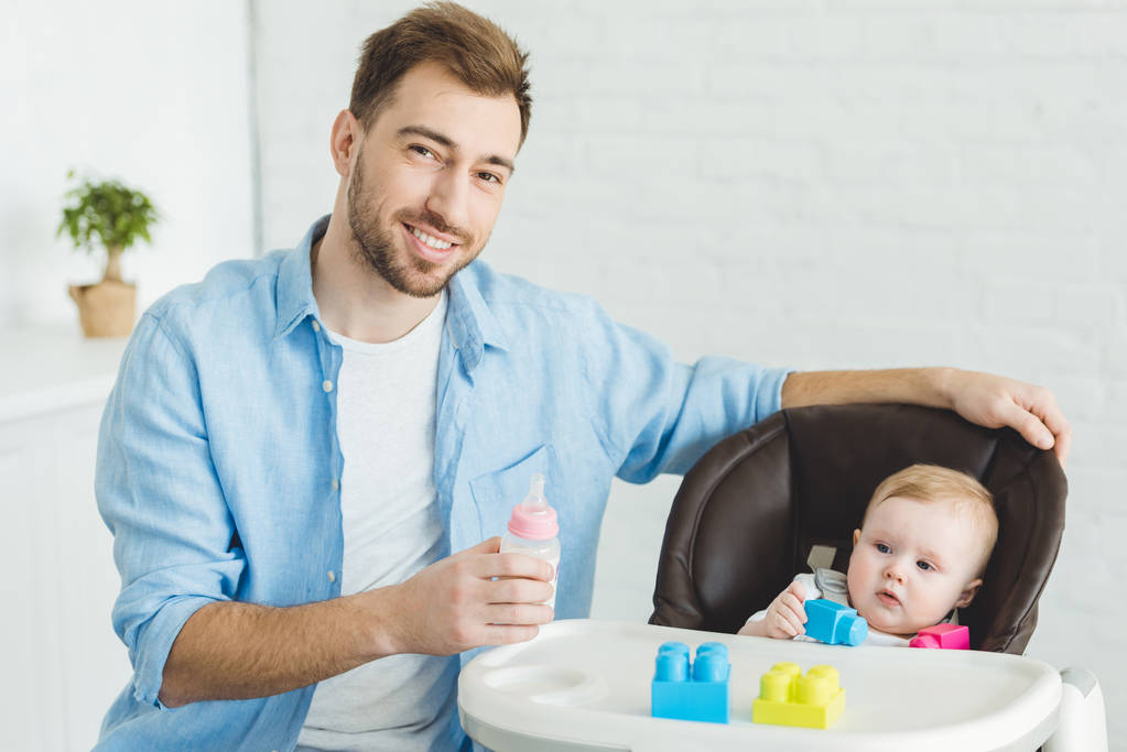 哺乳瓶と赤ちゃん椅子にプラスチック製のブロックで幼い娘を持つ父の笑みを浮かべてください ロイヤリティフリー写真 画像素材