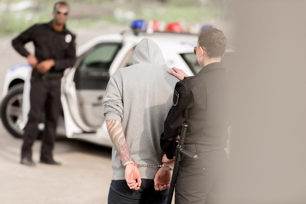 近くの車の後ろに立ってパートナー間手錠をかけられて男性犯罪者を逮捕した警官の背面図 ロイヤリティフリー写真 画像素材