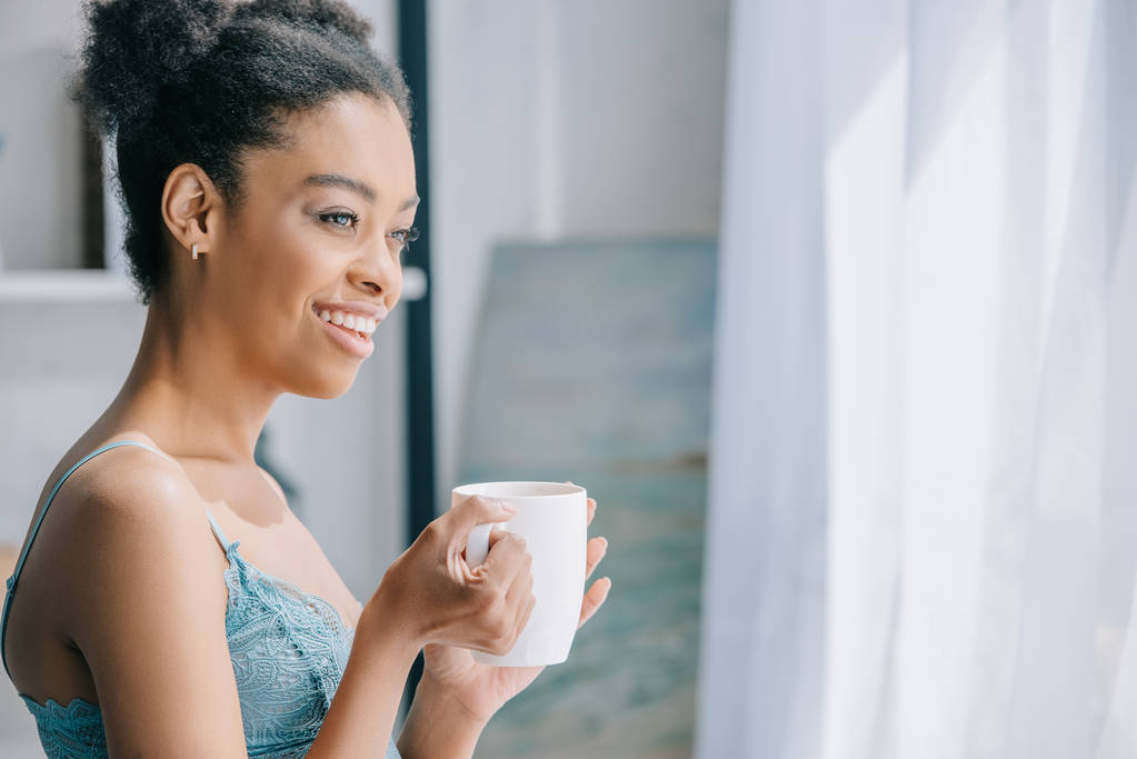 朝窓を見てコーヒーのカップと魅力的な微笑のアフリカ系アメリカ人女の子 ロイヤリティフリー写真 画像素材