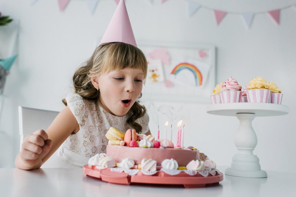 テーブルにバースデー ケーキのろうそくを吹き円錐形のかわいい子供 ロイヤリティフリー写真 画像素材