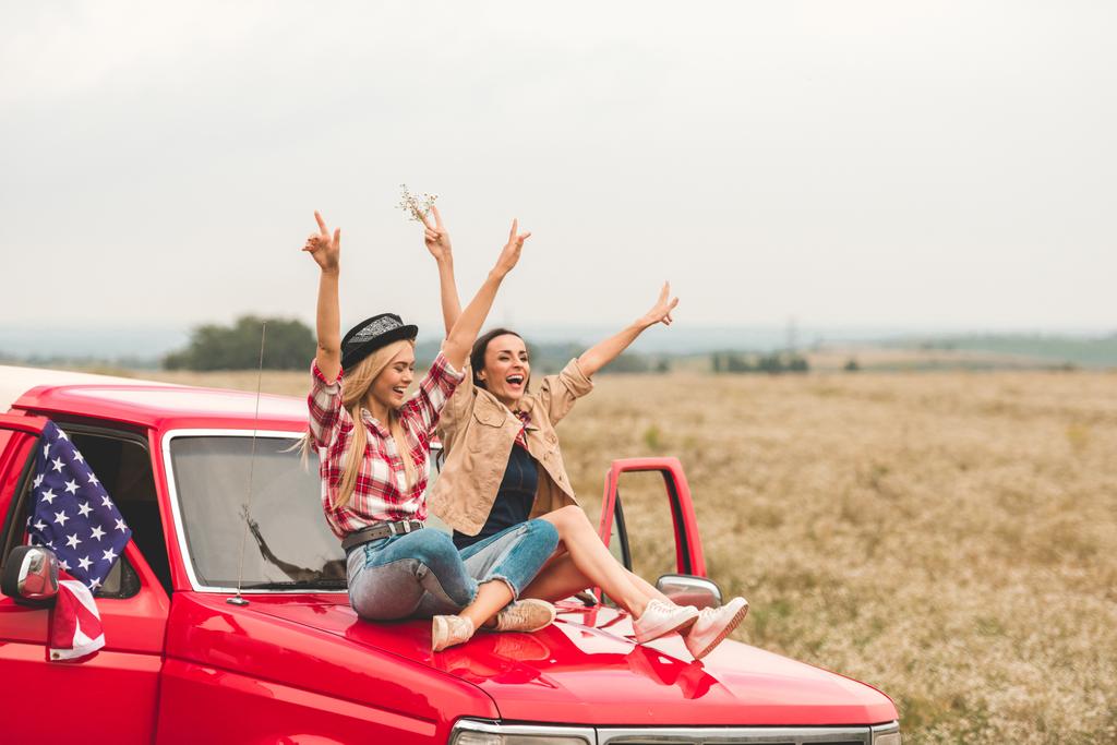 beautiful young girlfriends sitting on car hood with raised hands and showi...
