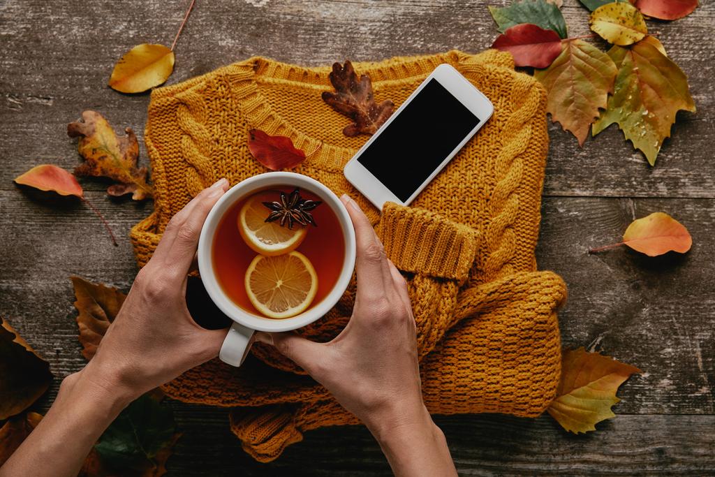 partial view of woman holding cup of tea on wooden tabletop with knitted sweater, smartphone and fallen leaves - Photo, Image