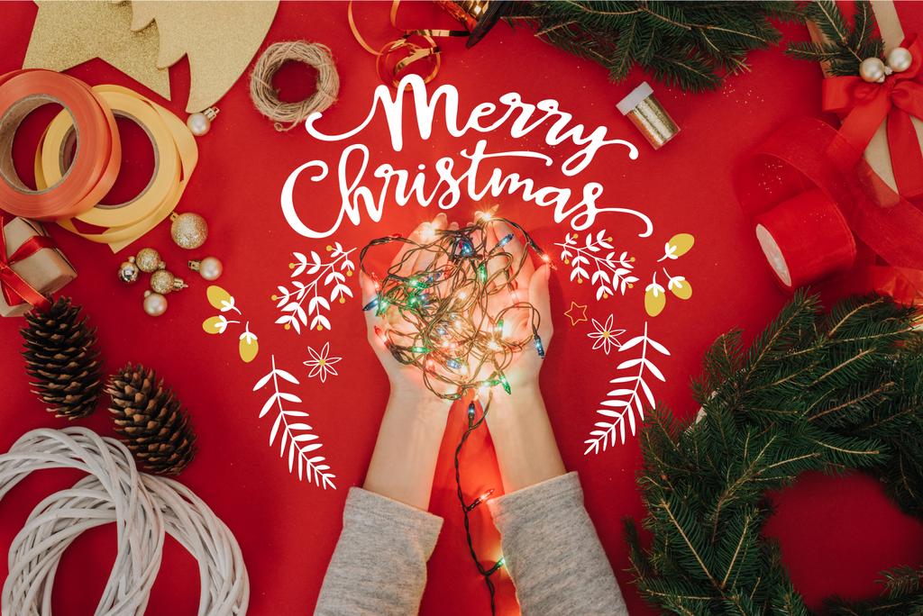 松の木の枝と メリー クリスマス の文字と赤い背景の手作りリース飾り手にクリスマス ライトを保持している女性のクロップ撮影 ロイヤリティフリー写真 画像素材