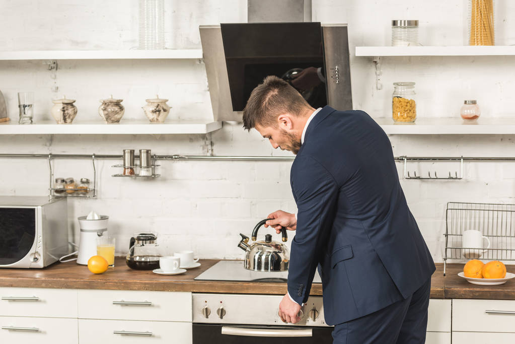キッチンで朝のストーブの上のやかんを入れてスーツを着たハンサムな男 ロイヤリティフリー写真 画像素材