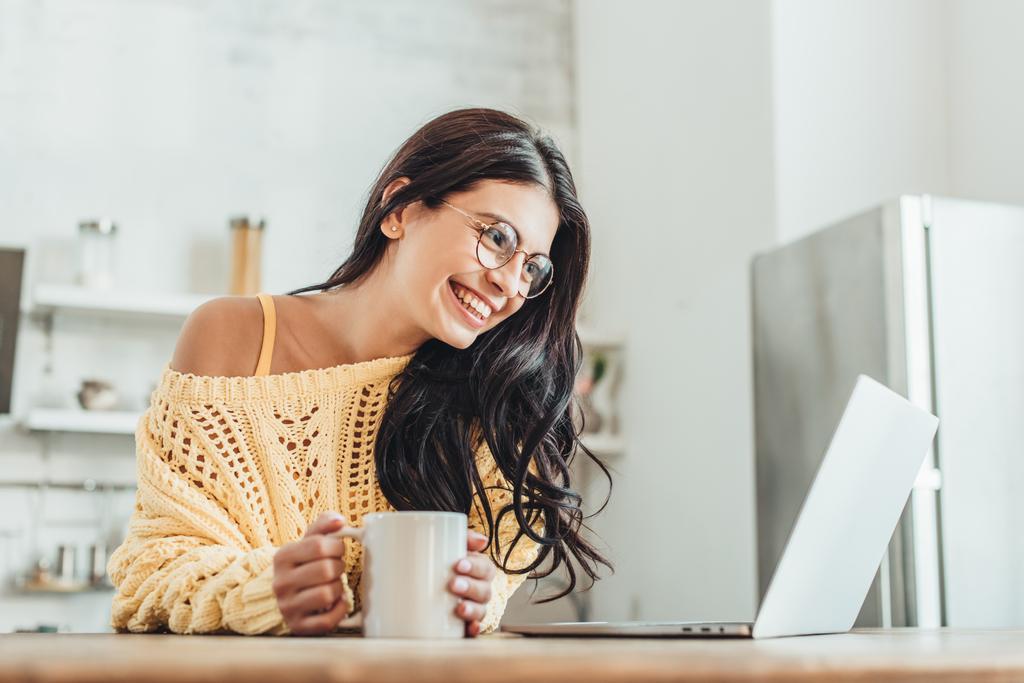 自宅の台所でノート パソコン コーヒー カップを持つ木製のテーブルに座って若い笑い女性フリーランサー ロイヤリティフリー写真 画像素材