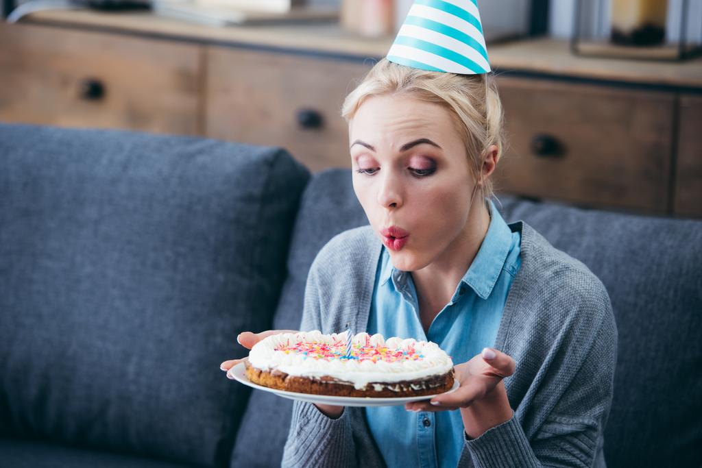 一人で自宅の誕生日を祝ってケーキの上のろうそくを吹いてパーティー帽子の女 ロイヤリティフリー写真 画像素材