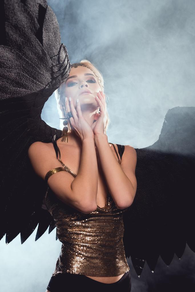 黒天使の羽と黄金アクセサリーの顔に触れると 煙のような背景でポーズ美しいセクシーな女性 ロイヤリティフリー写真 画像素材