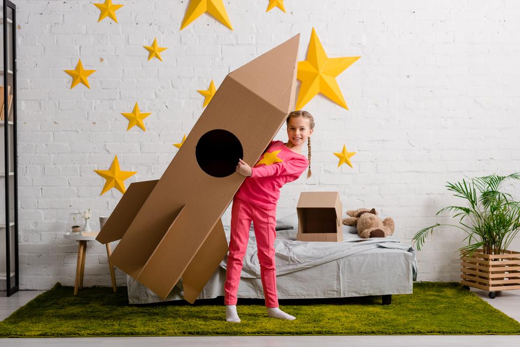 寝室に大きな段ボール ロケットを保持しているピンクの服で至福の子供 ロイヤリティフリー写真 画像素材