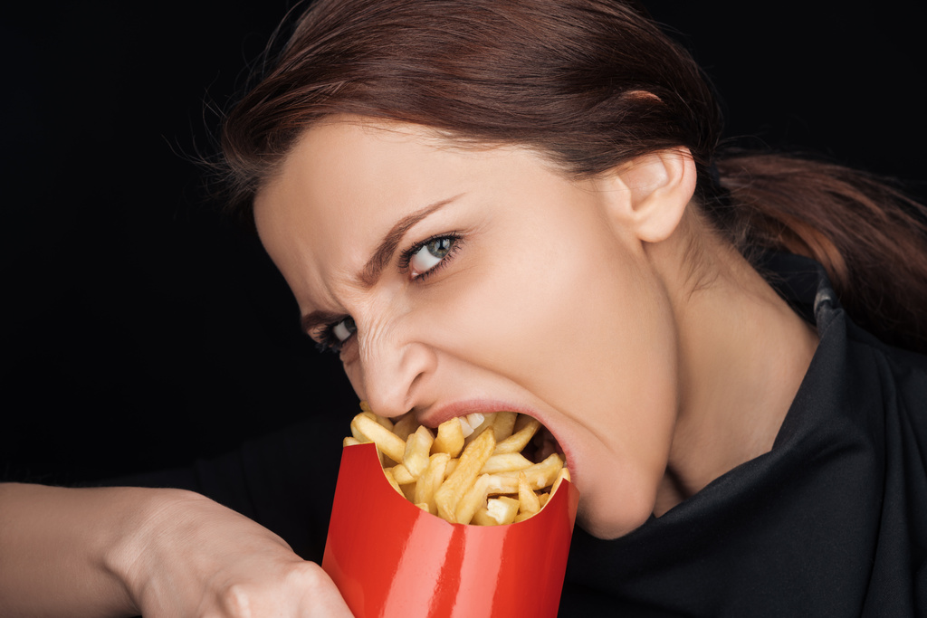 emotional woman eating salty french fries while looking at camera isolated ...