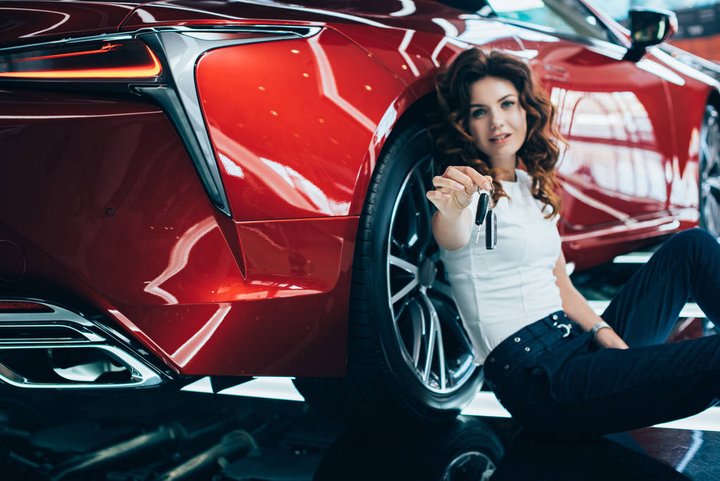 新しい赤い車の近くの床に座っている幸せな女性とキーを保持 ロイヤリティフリー写真 画像素材