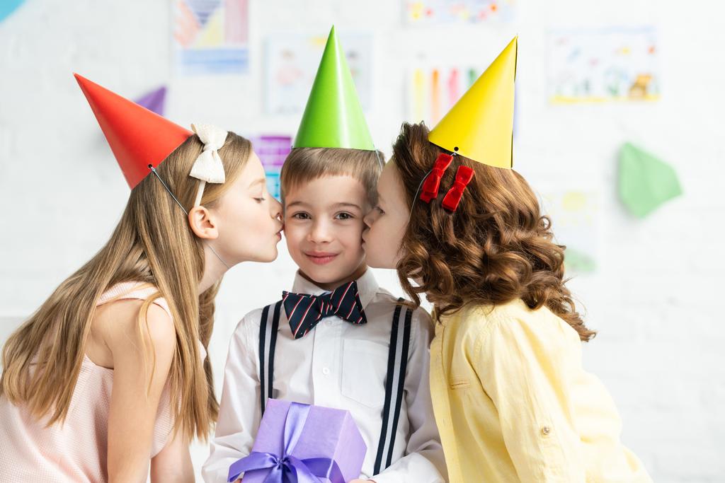 パーティーキャップの子供たちは 自宅で誕生日パーティー中にギフトボックスで頬の愛らしい男の子にキス ロイヤリティフリー写真 画像素材