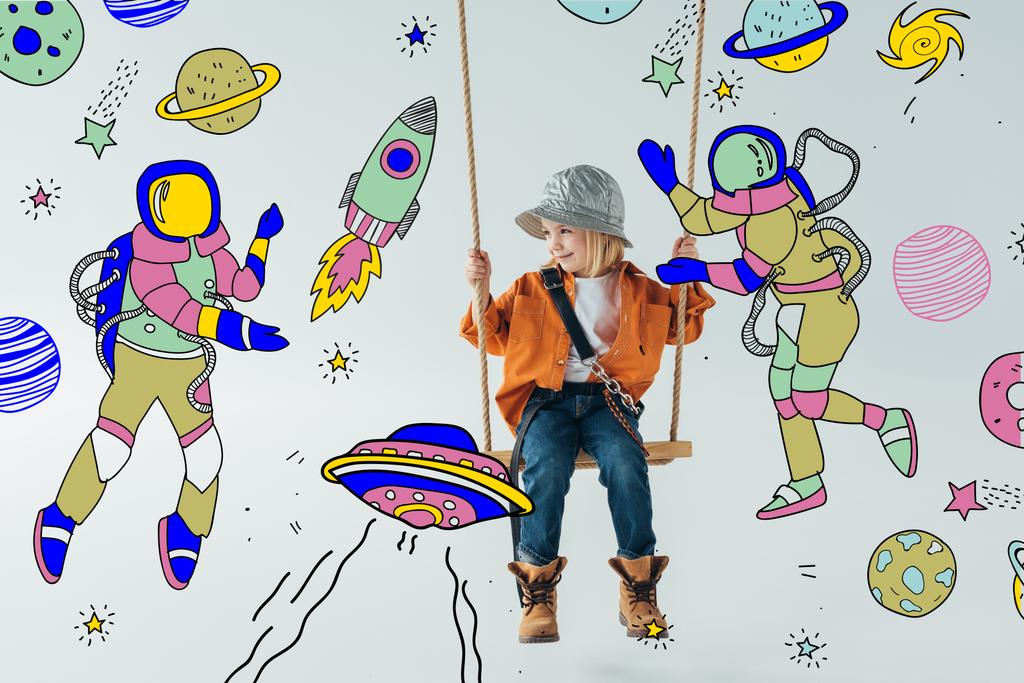ジーンズとオレンジ色のシャツのかわいい子供は ブランコに座って 灰色の背景に宇宙飛行士のイラストと妖精の空間を見て ロイヤリティフリー写真 画像素材