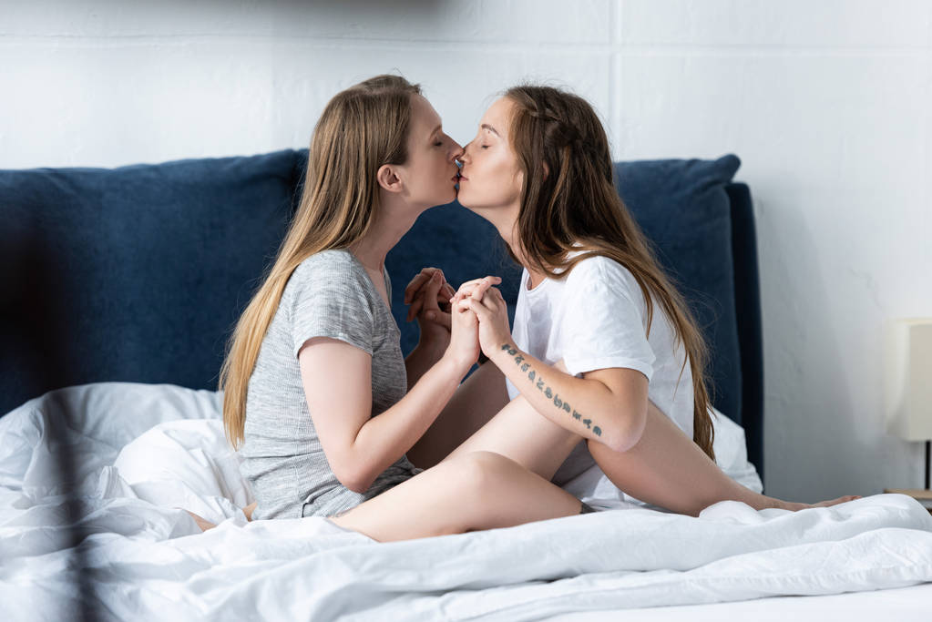 Widok z boku dwóch lesbijek trzymając ręce i całuje na łóżku - Zdjęcie, obr...