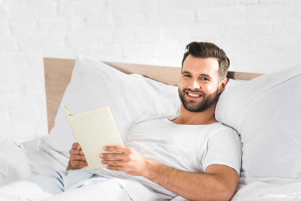 Foto e imagen de stock sin derechos de Guapo feliz hombre acostado en la  cama y leyendo libro en la mañana