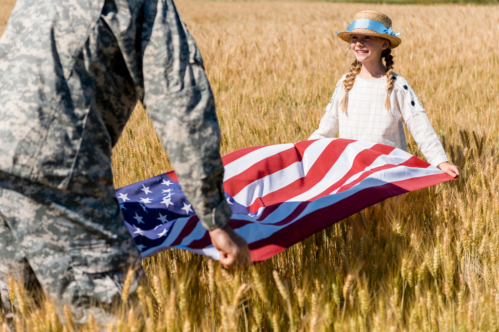 フィールドでかわいい娘とアメリカの旗を保持する軍服を着た男のトリミングビュー ロイヤリティフリー写真 画像素材