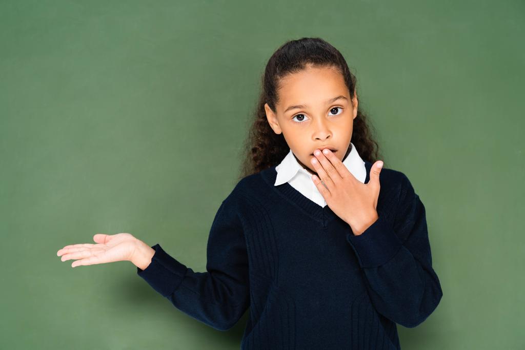 緑の黒板の近くに立っている間 手で口を覆い 手で指差す衝撃を受けたアフリカ系アメリカ人の女子学生 ロイヤリティフリー写真 画像素材
