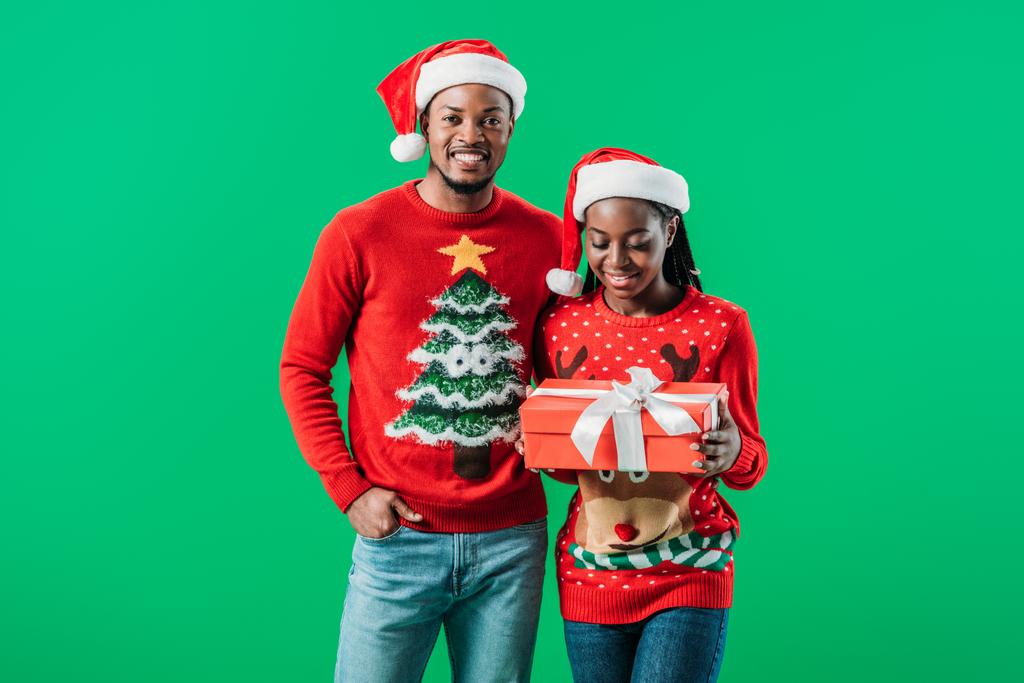 赤いクリスマスセーターとサンタ帽子のアフリカ系アメリカ人男性は 緑の上に手で隔離されたギフトボックスを保持している女性の近くに ロイヤリティフリー写真 画像素材