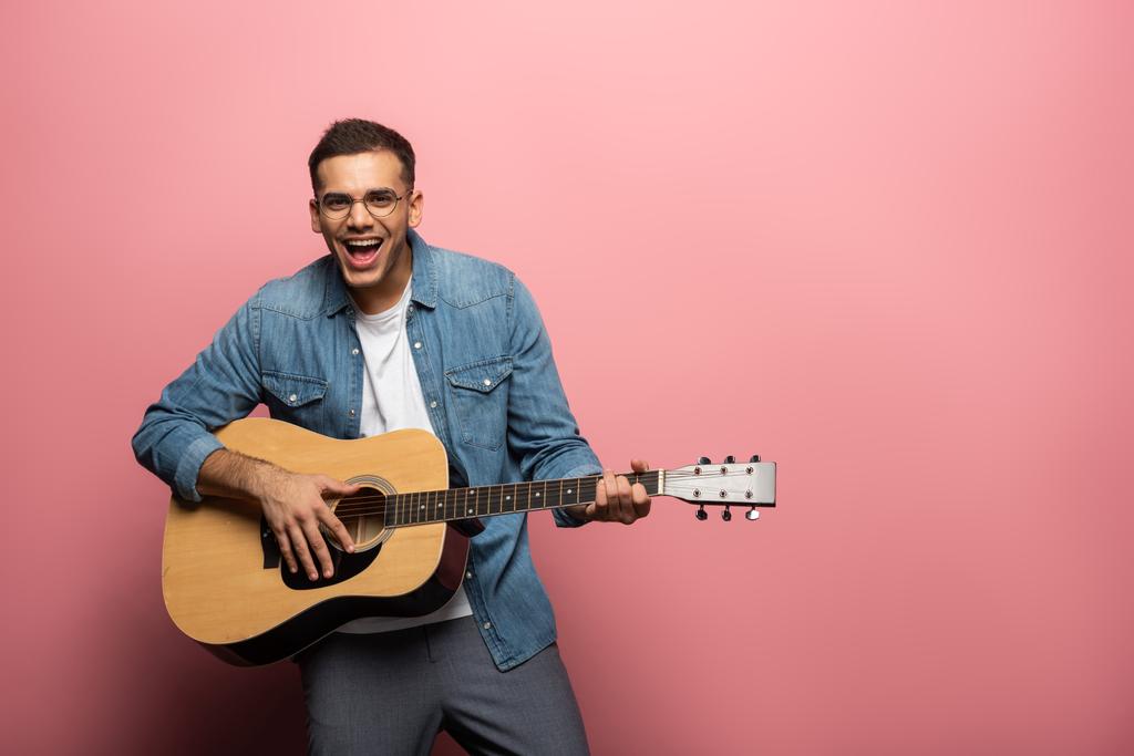 若い笑い男はピンクの背景にアコースティックギターを演奏しながらカメラを見て ロイヤリティフリー写真 画像素材