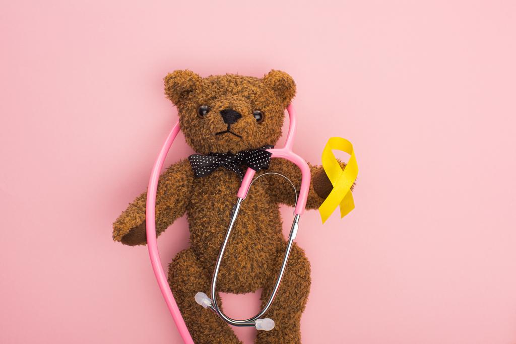 ピンクの背景に茶色のテディベアに黄色のリボンと聴診器のトップビュー 国際小児がんの日の概念 ロイヤリティフリー写真 画像素材