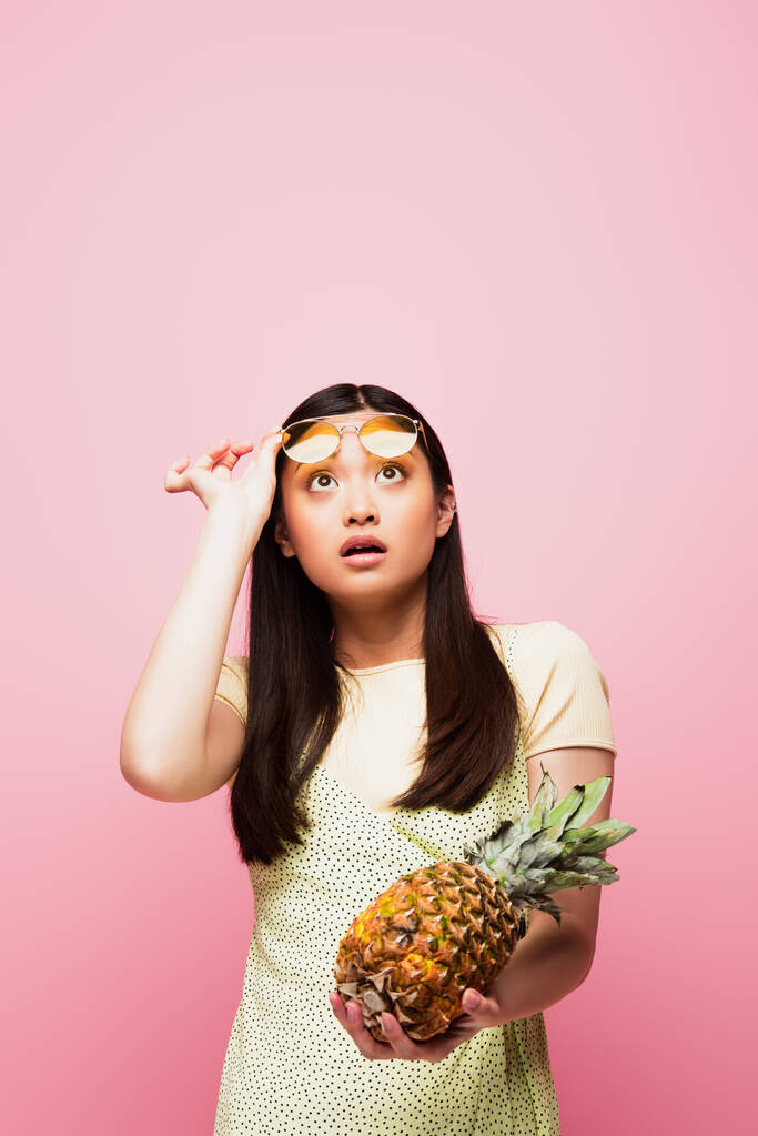 ピンク色に隔離された新鮮なパイナップルを持ち上げるサングラス姿のアジア系の女の子 ロイヤリティフリー写真 画像素材