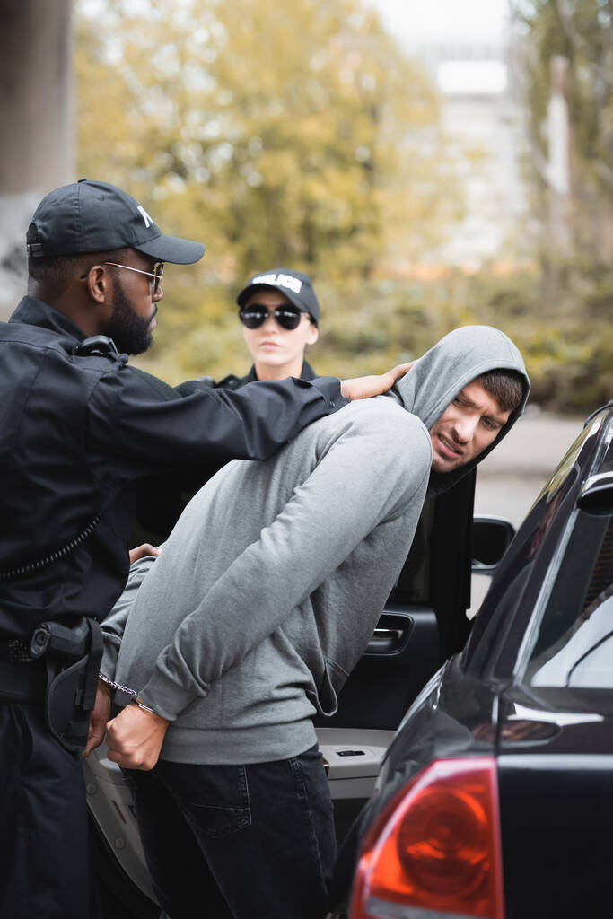 アフリカ系アメリカ人警官が同僚の近くで怒ったフード犯罪者を逮捕し屋外で車をパトロール ロイヤリティフリー写真 画像素材