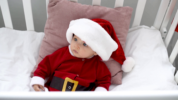びっくりかわいい赤ちゃんサンタ衣装よそ見やベビーベッド 自宅で座っているクリスマスのコンセプトで ストック動画映像