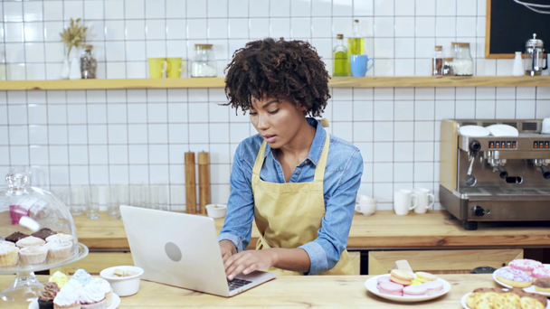 美しい笑顔のカフェでデザートをカウンターの後ろにノート パソコンを使用してアフリカ系アメリカ人の女性レジ係 ストック動画映像