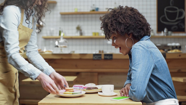 美しいドーナツ ケーキ マカロンをもたらす女性ウェイトレス笑顔カフェでアフリカ系アメリカ人の女性客 ストック動画映像