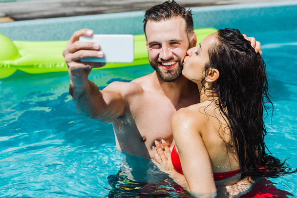 スイミングプールで自分撮りを取る幸せな男性の頬にキス女性の選択的な焦点 ロイヤリティフリー写真 画像素材
