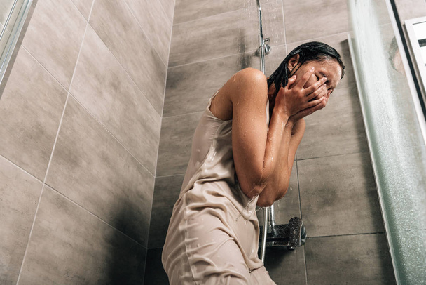 depressed woman covering face with hands while crying in shower at home - P...