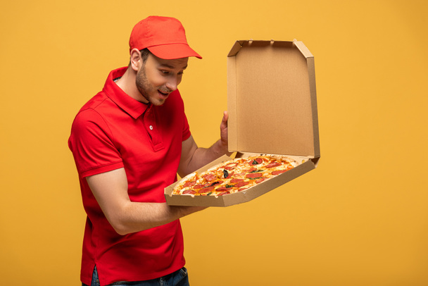 Happy delivery man in red uniform looking at box with delicious pizza isola...