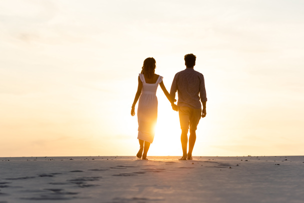 Gün batımında plajda güneşe karşı yürürken el ele tutuşan kadın ve erkeğin  siluetleri. Telifsiz Fotoğraf ve Stok Görsel