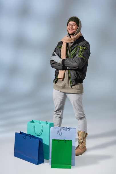 灰色の買い物袋の近くに腕を組んで立っている幸せな男の完全な長さ ロイヤリティフリー写真 画像素材